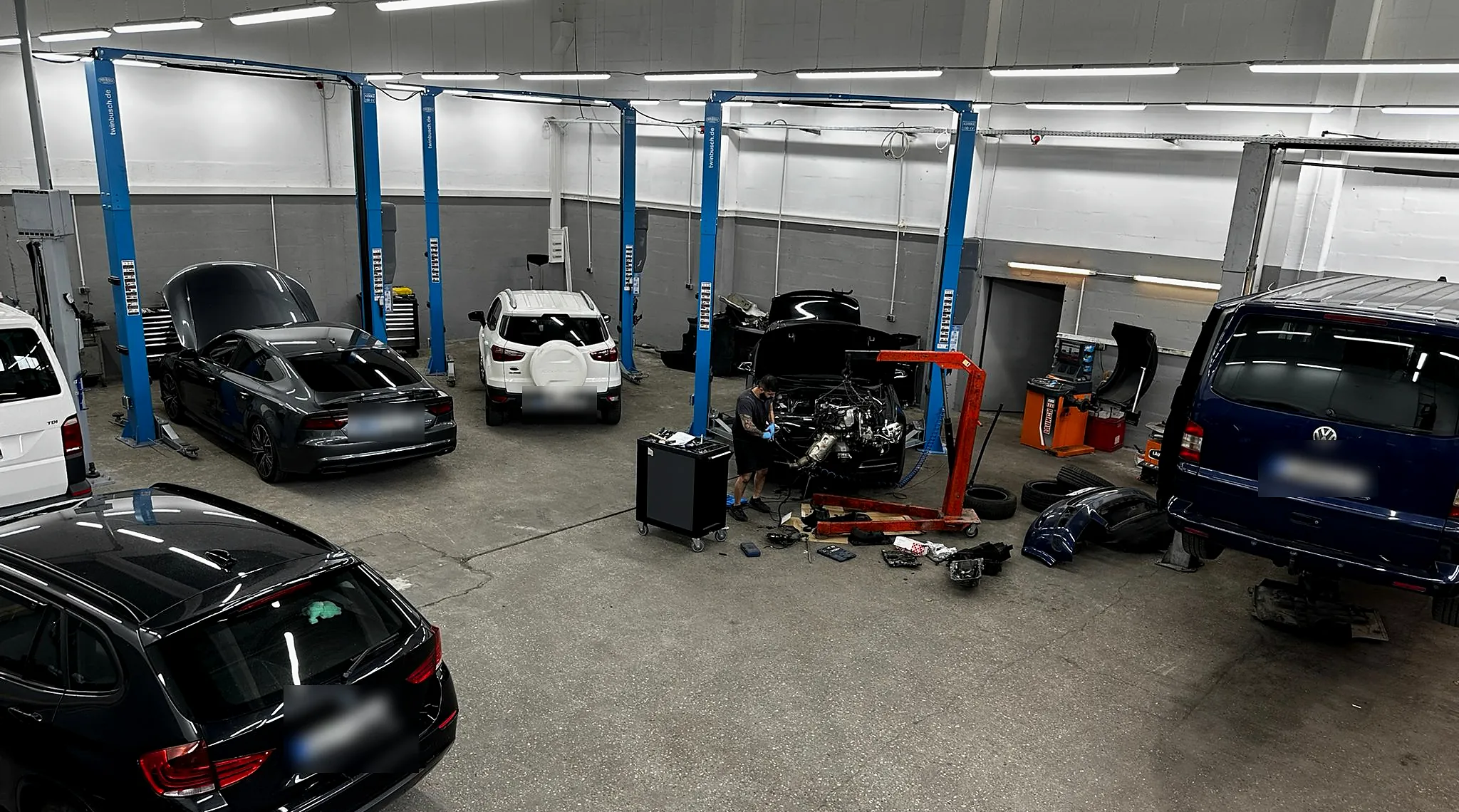Blick in die Werkstatthalle mit mehreren Fahrzeugen an Hebebühnen, Werkzeugwagen und Ersatzteilen